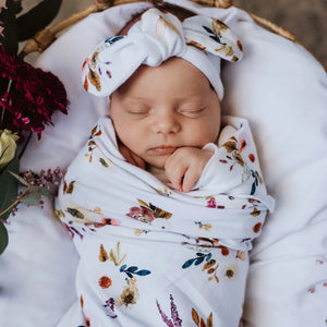 Newborn baby swaddled in a floral blanket with a matching headband, surrounded by flowers.