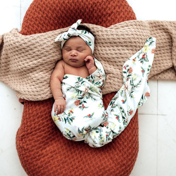 Newborn baby wrapped in a floral blanket with a knitted brown blanket and headband on a white background